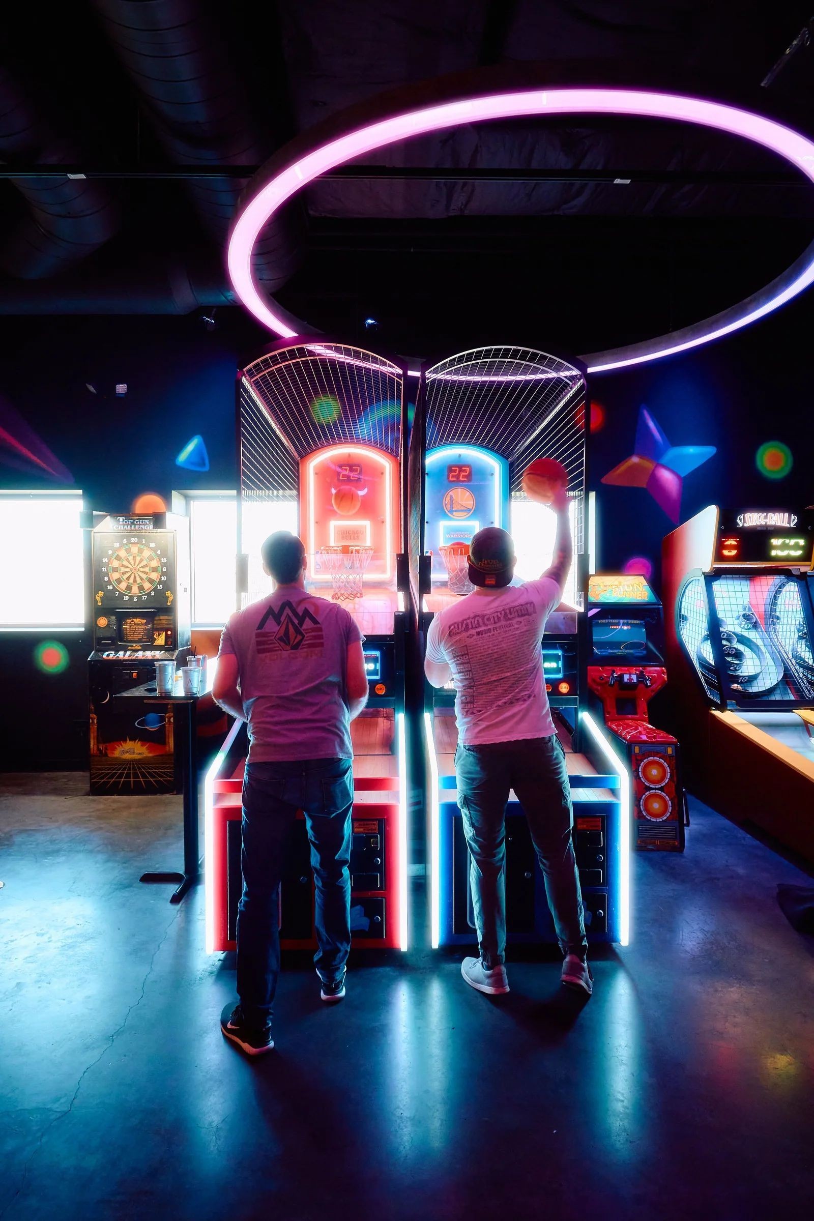Two men playing an arcade basketball hoop game side by side under a glowing neon ring at Asylum Bar + Arcade