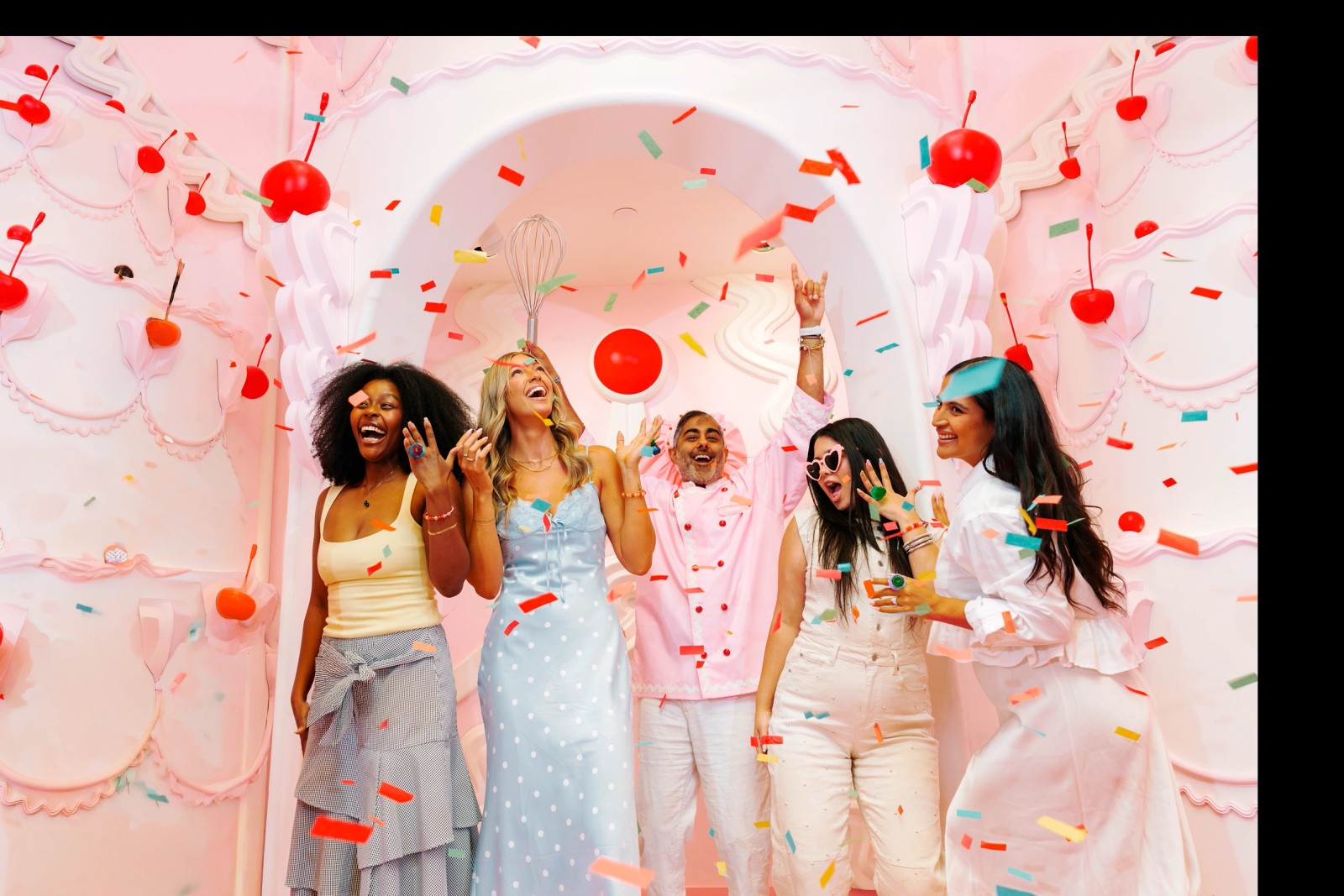 A group of visitors cheering under a shower of confetti inside the Museum of Ice Cream's pink cake-themed chapel room, decorated with sculpted frosting and cherries