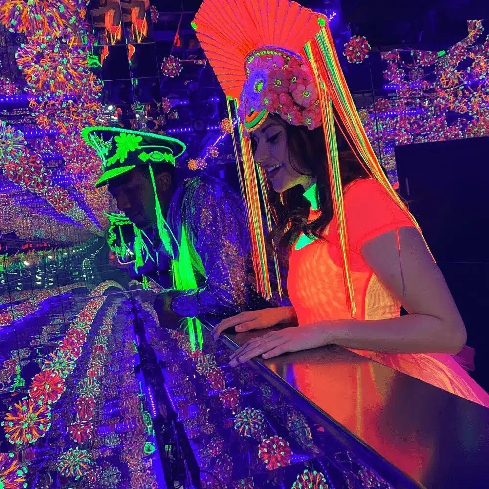 Two visitors in glowing UV face paint and neon headdresses lean over a mirrored rail inside one of Wink World's infinity mirror rooms