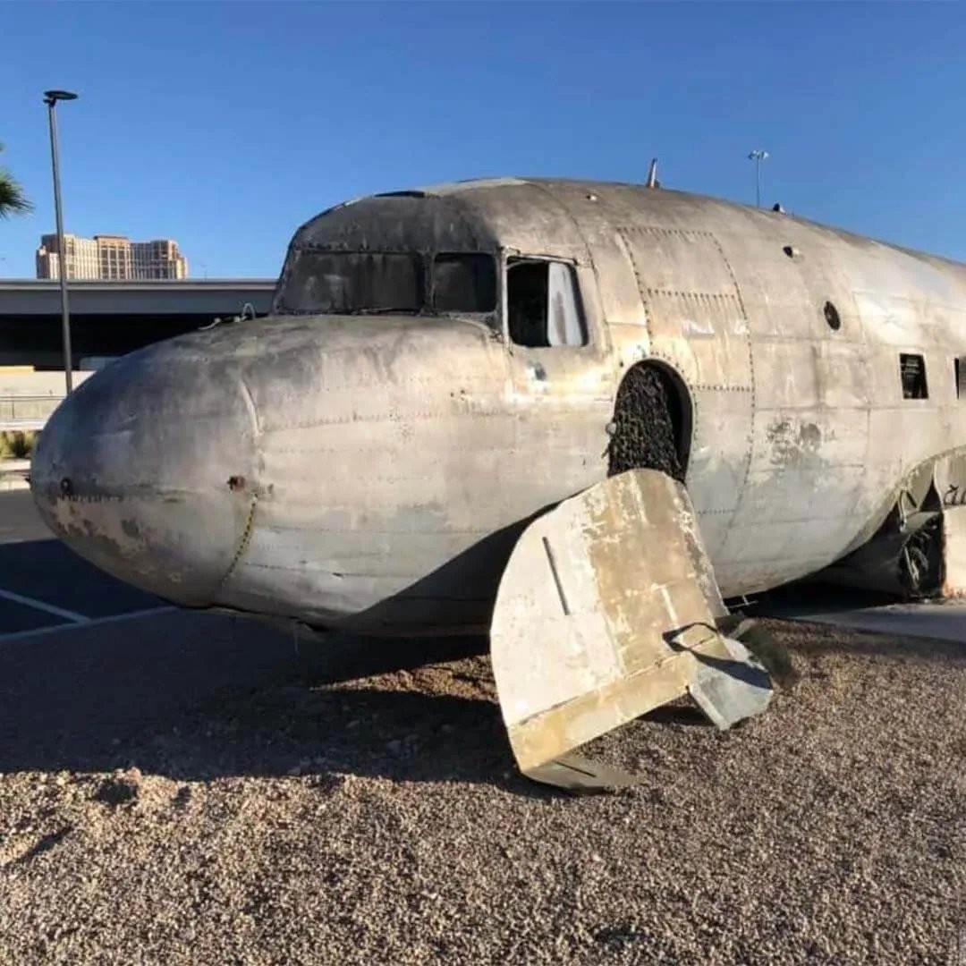 The weathered nose and cockpit of a real WWII-era C-47 Cargo Plane parked outdoors on Art Island, with a Las Vegas parking garage visible behind it in daylight