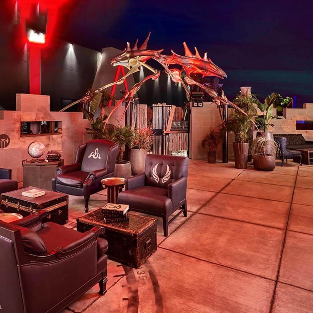 Gilly, a 21-foot-long, 16-foot-tall stainless steel alien-like sculpture, arching over an outdoor lounge area at AREA15 at night, lit in red and gold