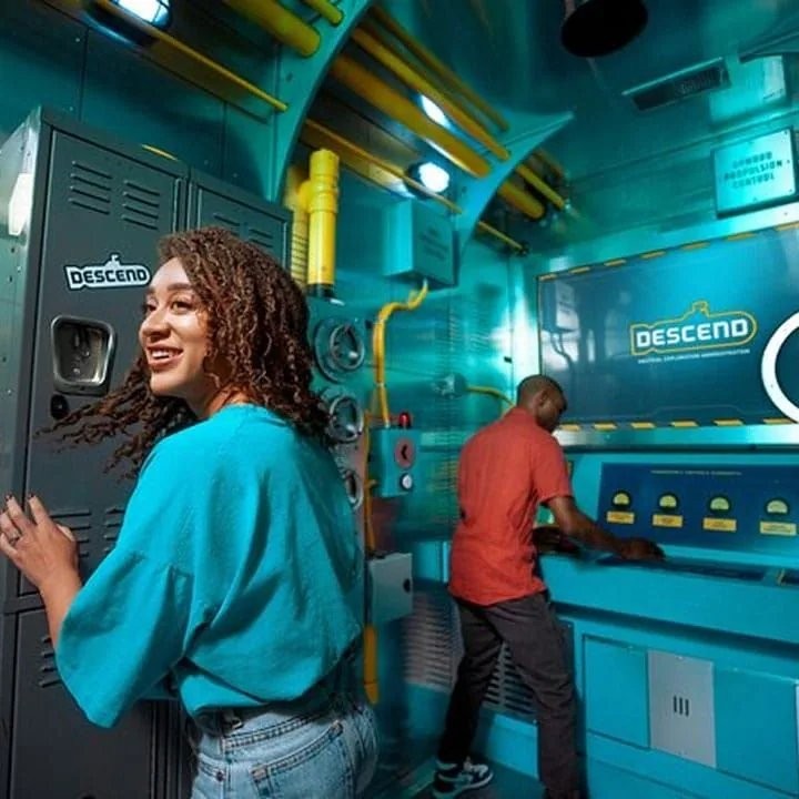 Two players inside The Escape Game's 'Descend' room, a teal-lit submarine-themed set with control panels and lockers