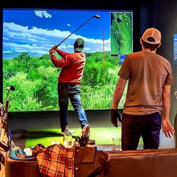 A golfer swinging at an indoor golf simulator bay at Five Iron Golf while another guest watches, golf bags and gear in the foreground