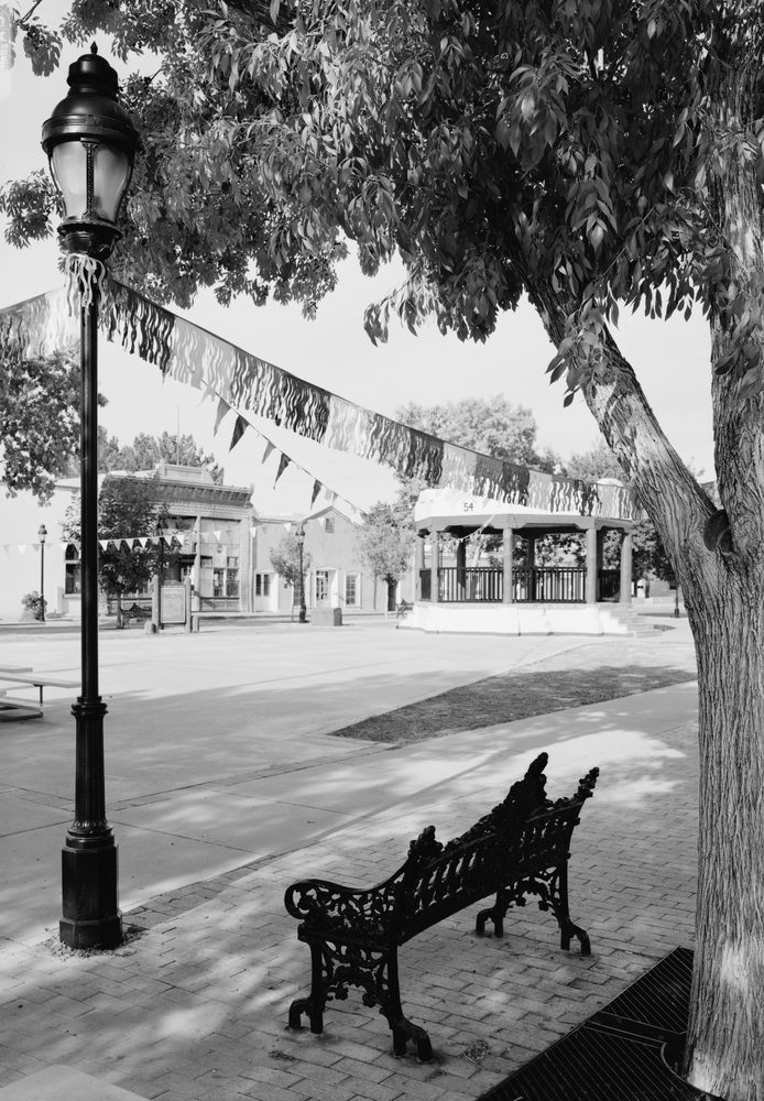 A black-and-white photo of the historic Mesilla Plaza in Mesilla, New Mexico, showing a wrought-iron lamppost, a park bench, bunting, and the plaza's gazebo and surrounding adobe storefronts