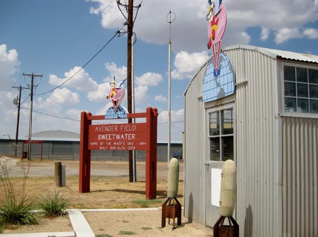 The Avenger Field entrance sign in Sweetwater, Texas, topped with the Fifinella WASP mascot, in front of a corrugated-metal hangar building