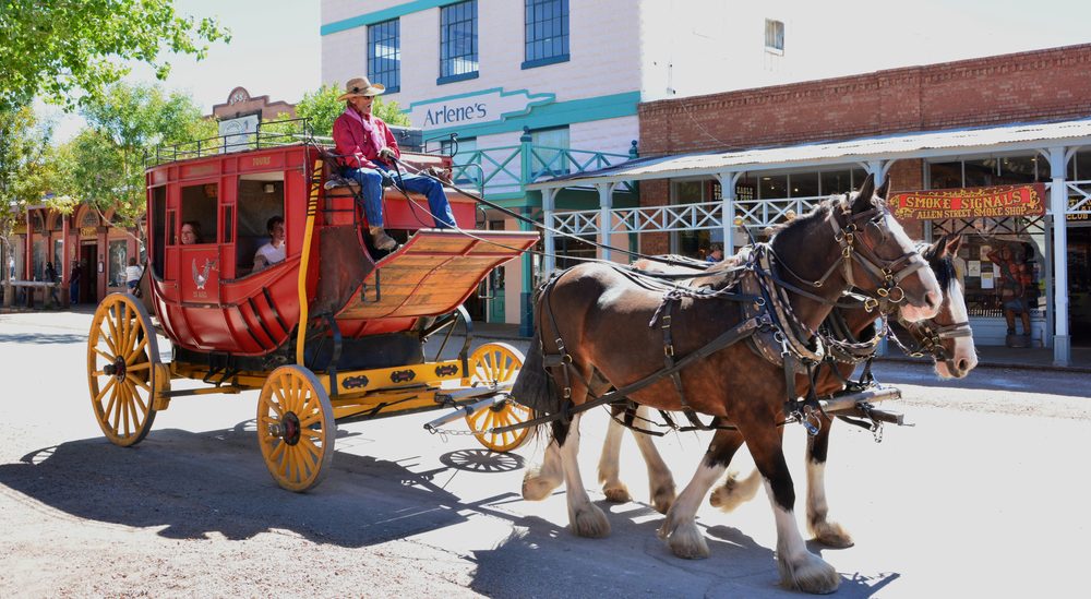 A stagecoach pulled by two Clydesdale horses driving down Allen Street in Tombstone, Arizona, past storefronts including a smoke shop and a general store