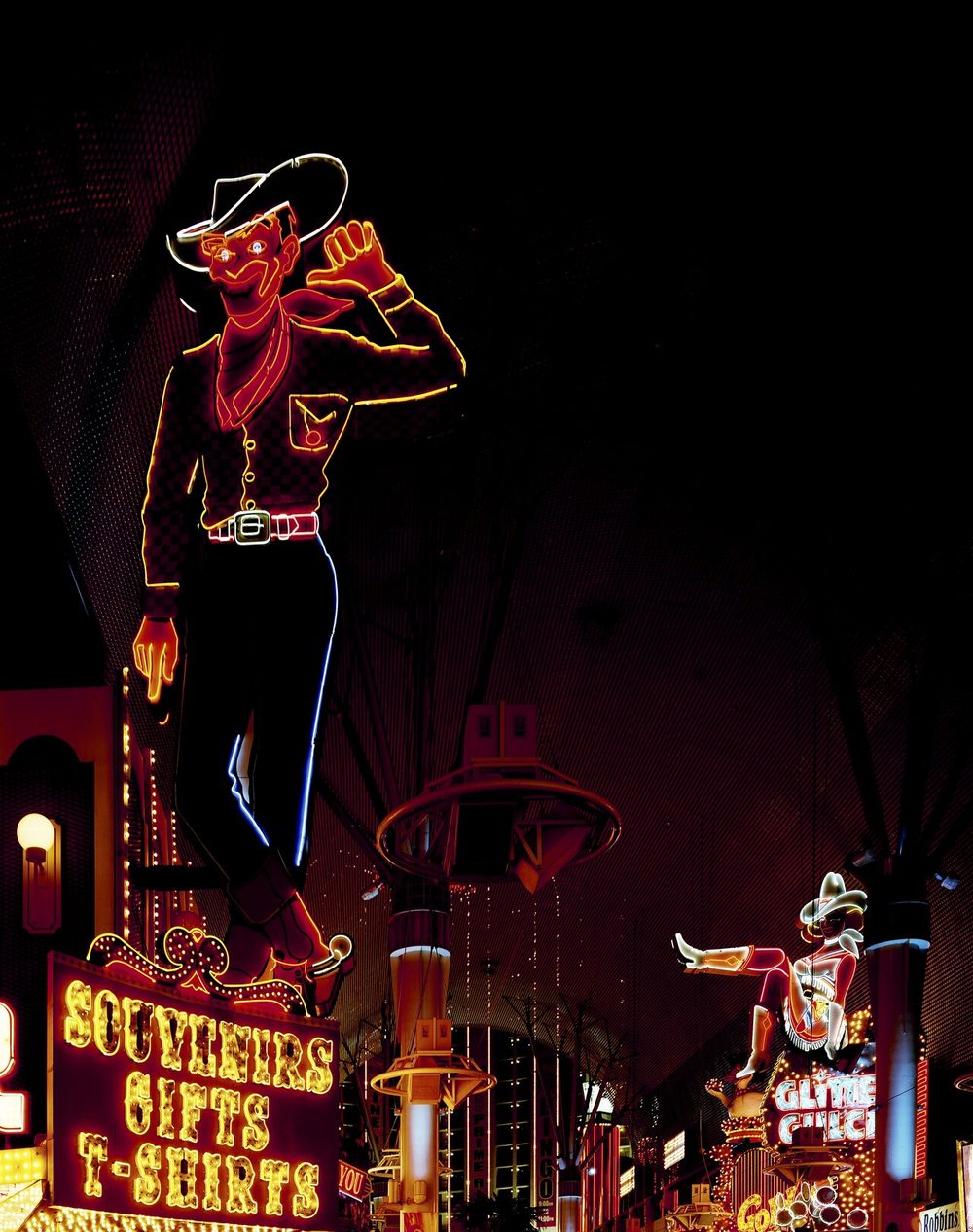 Vegas Vic, a 40-foot neon cowboy sign with a waving arm, mounted above a souvenir shop on Fremont Street, with Vegas Vickie's neon cowgirl sign visible in the distance