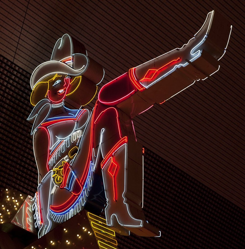 Close-up of Vegas Vickie, a neon cowgirl sign with one leg kicked outward, wearing a fringed outfit and cowboy hat