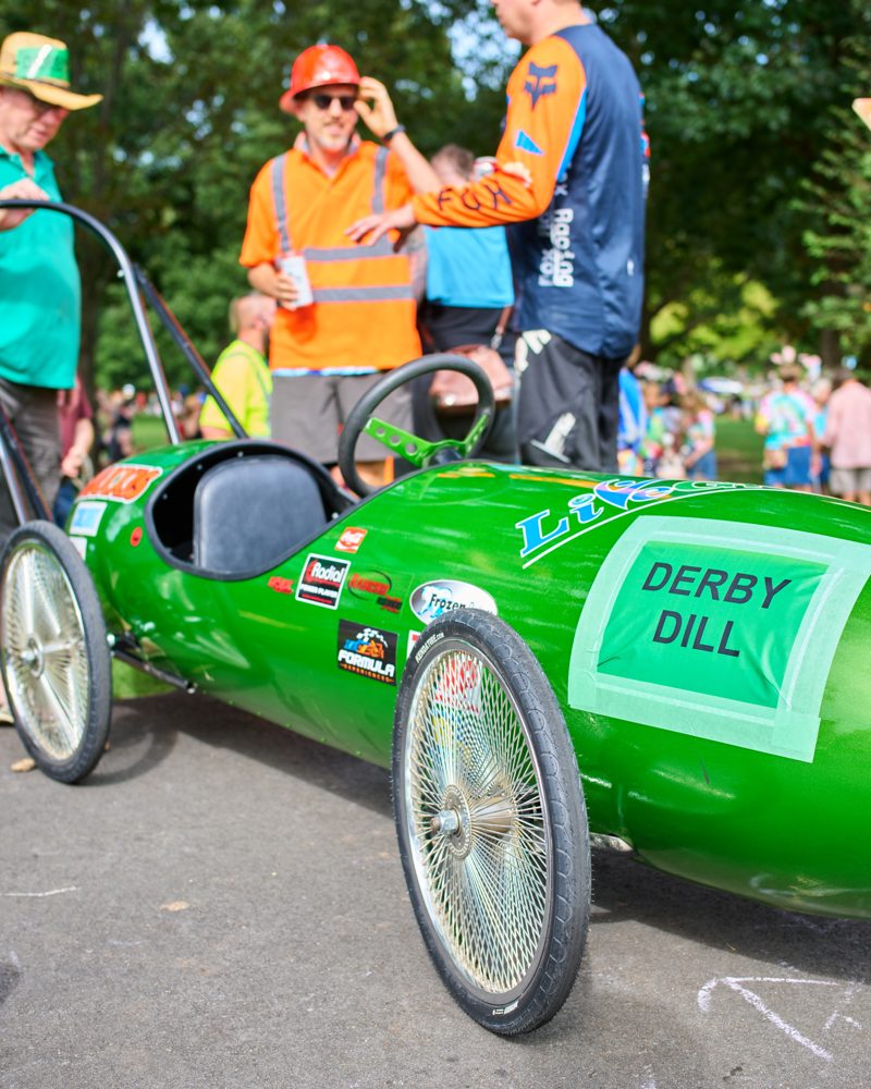 A past entrant's handmade green soapbox car labeled DERBY DILL with racing decals, with costumed entrants nearby