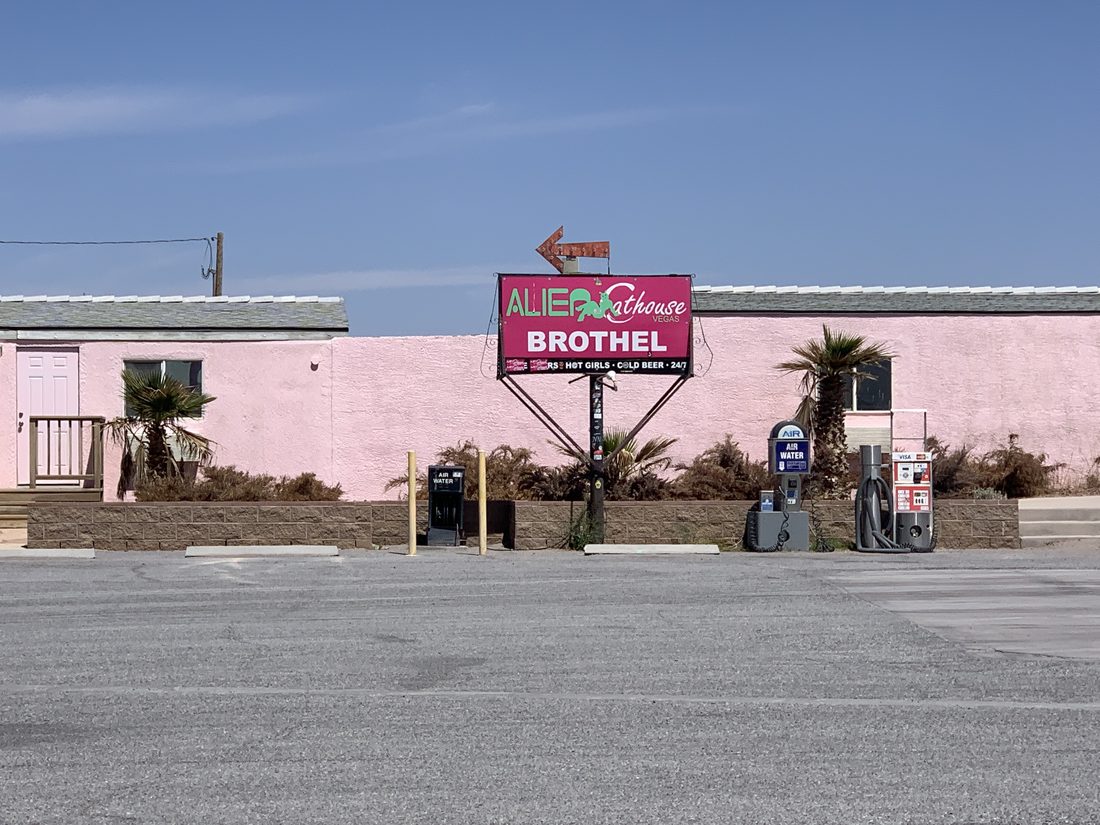 The pink stucco exterior of the Alien Cathouse brothel in Amargosa Valley, Nevada, with a lit roadside sign reading Alien Cathouse Brothel next to gas pumps on US-95