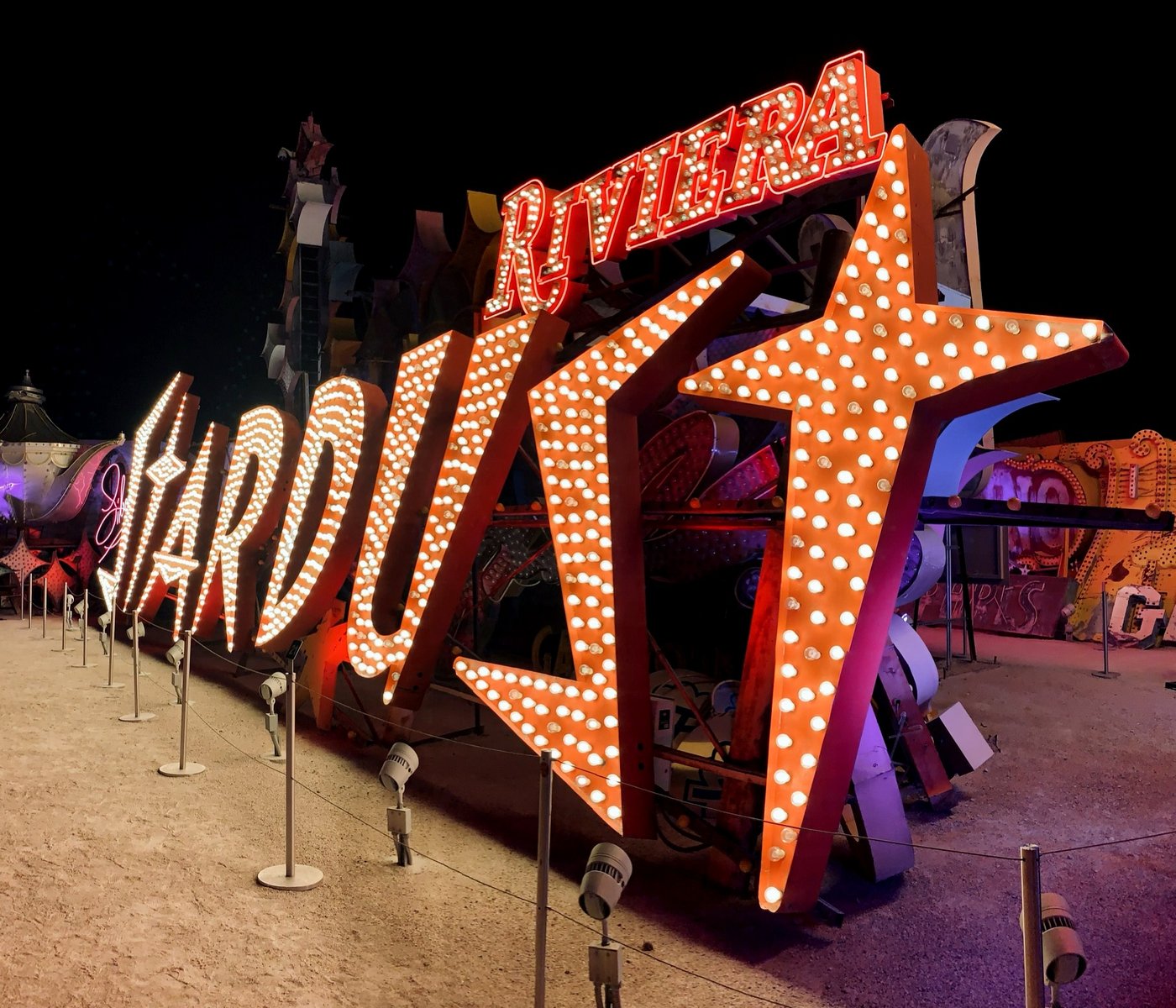 The restored Stardust sign at the Neon Museum's Neon Boneyard at night, showing a large illuminated orange starburst above the word Stardust, with the Riviera sign visible behind it