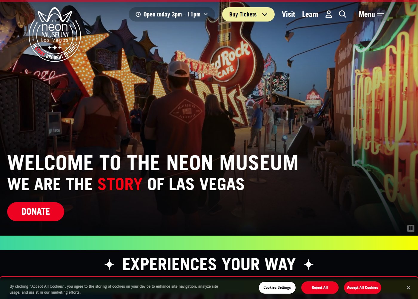 The Neon Museum's own homepage, showing visitors walking through the Neon Boneyard at dusk past a giant Hard Rock Cafe guitar sign and the restored Stardust letters