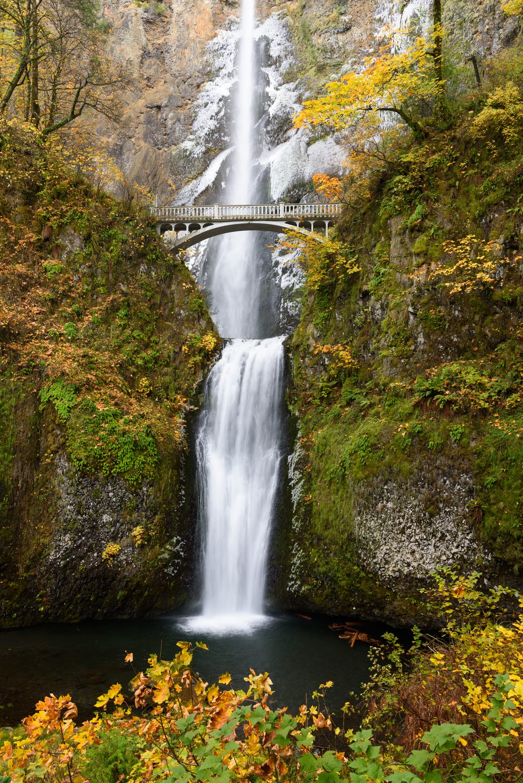 Multnomah Falls, a tall two-tiered waterfall in the Columbia River Gorge, with a stone footbridge crossing between the upper and lower tiers, surrounded by autumn foliage