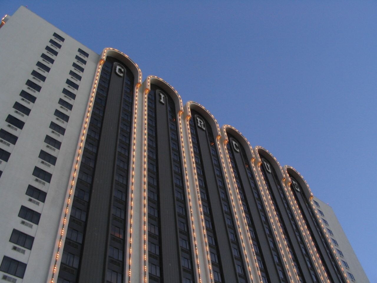A low-angle view up the side of Circus Circus Reno's hotel tower, with illuminated marquee letters spelling out C-I-R-C-U-S running up the building's arched columns against a clear blue sky