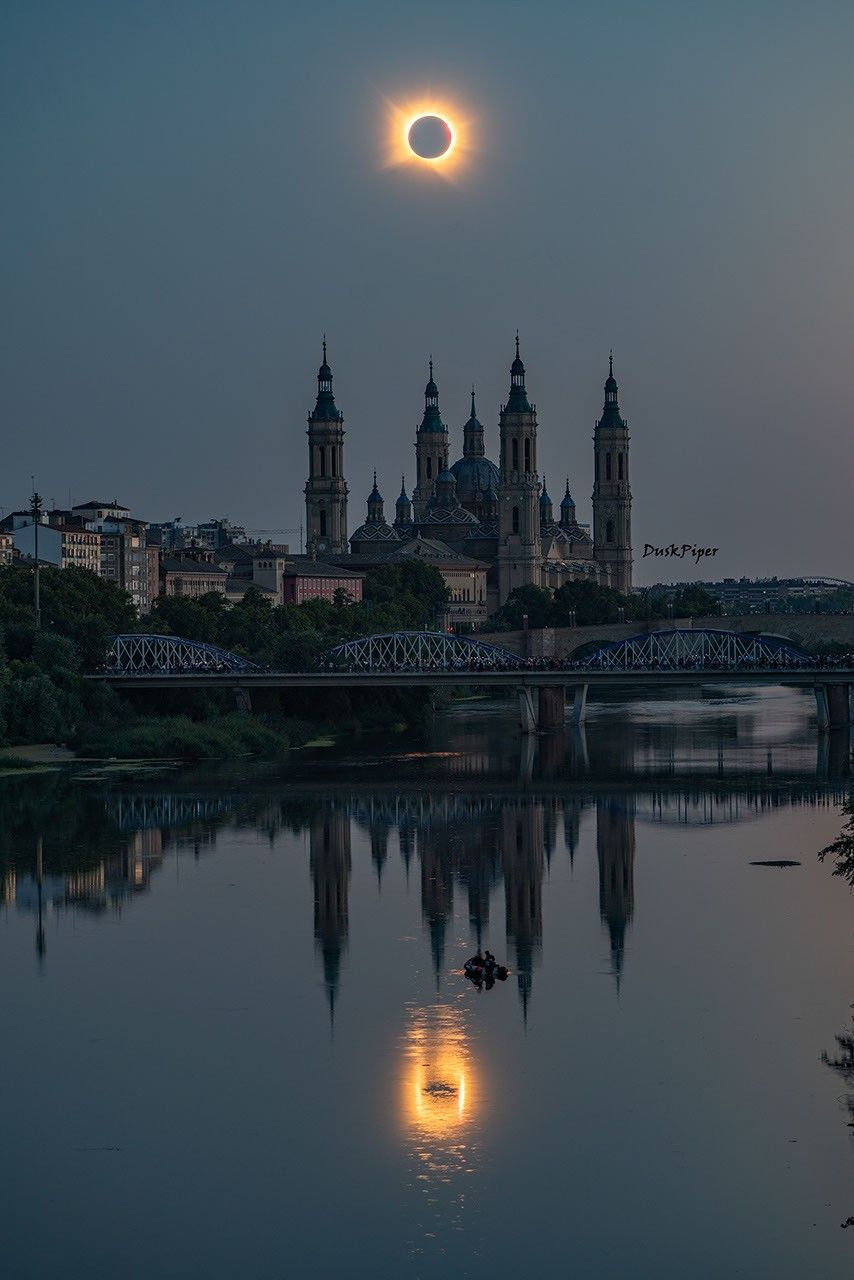 Two views of totality during the August 12, 2026 total solar eclipse over Zaragoza, Spain, one over the Cathedral-Basilica of Our Lady of the Pillar and one reflected in the Ebro River with people boating below