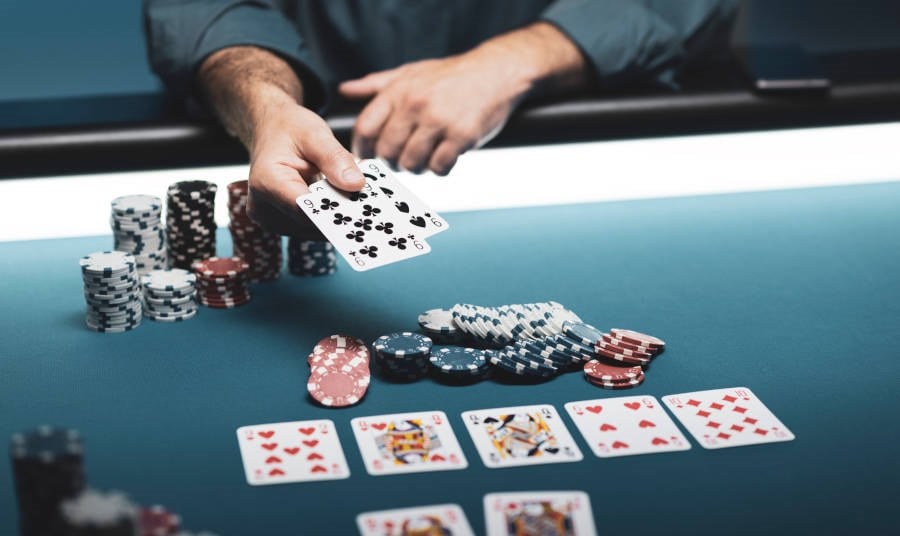 A real photo of a hand dealing cards over stacked casino chips with five community cards spread on an Ultimate Texas Hold'em table