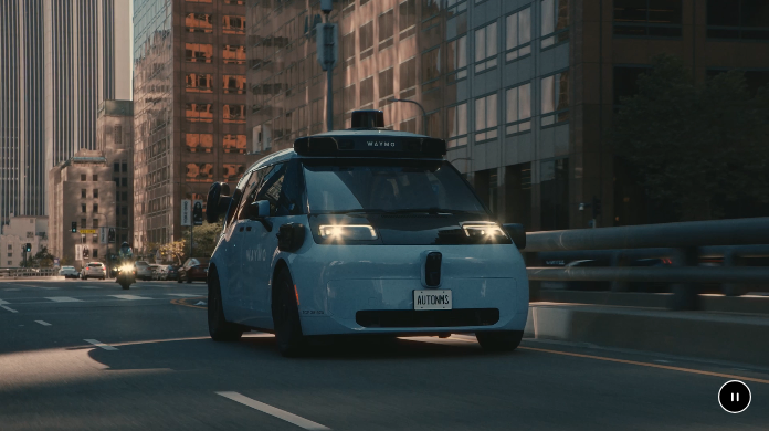 A real Waymo autonomous vehicle, roof-mounted sensor visible, driving through a city street at dusk, from Waymo's official company blog