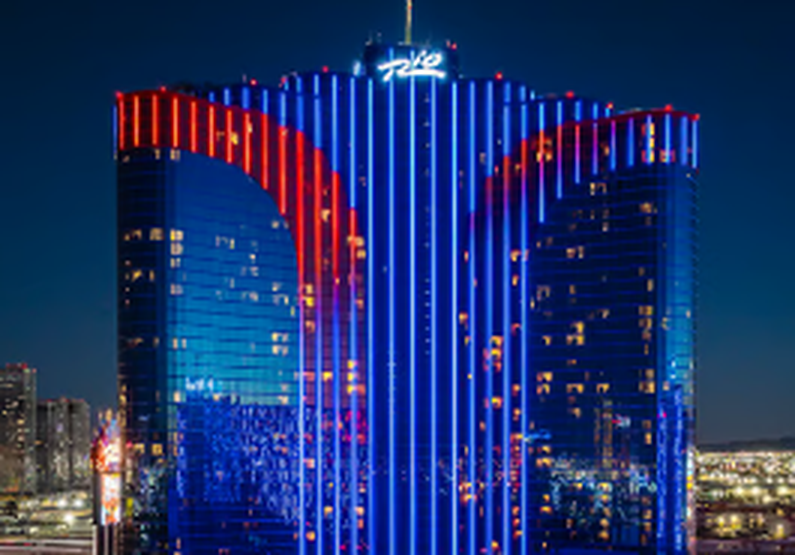 The Rio Las Vegas's official site showing the hotel's twin towers lit up in blue and red neon at night, with the renovated pool deck visible below